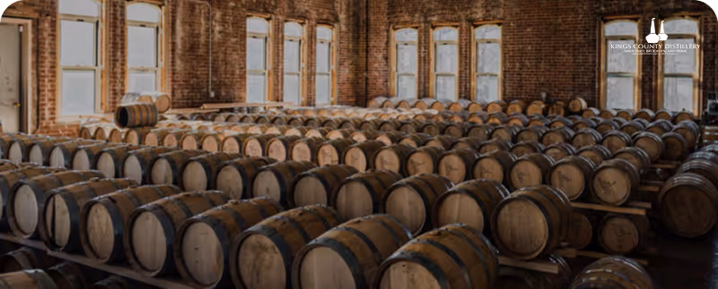 Rows of wooden barrels aging inside a distillery warehouse with brick walls and tall windows.