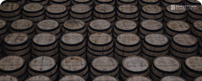 Rows of wooden barrels stacked, each marked with Bardstown Bourbon Company logo and text on top.