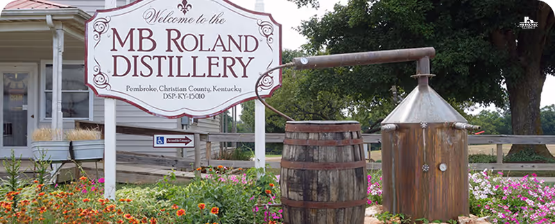 Sign reading MB Roland Distillery in Kentucky with wooden barrel and copper still in garden flowers.