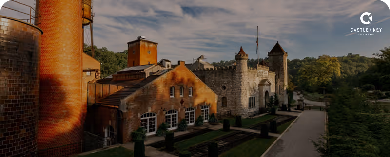 Castle & Key Distillery with brick buildings, stone towers, and surrounding greenery under a partly cloudy sky.