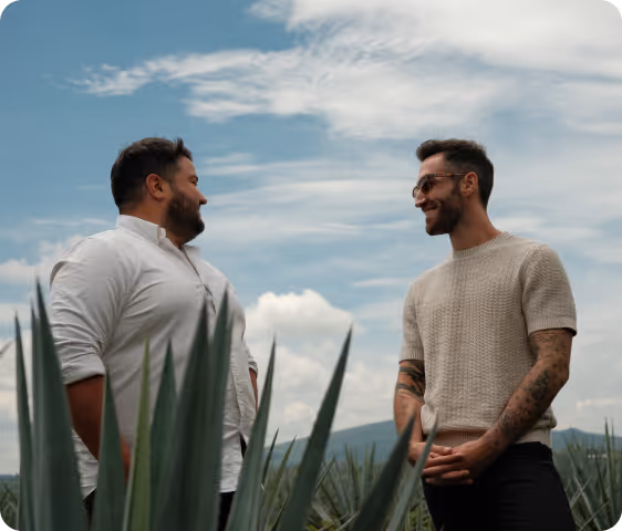 Two men smiling and talking outdoors with a blue sky and green plants in the foreground.