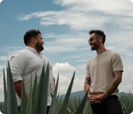 Two men smiling and talking outdoors with a blue sky and green plants in the foreground.