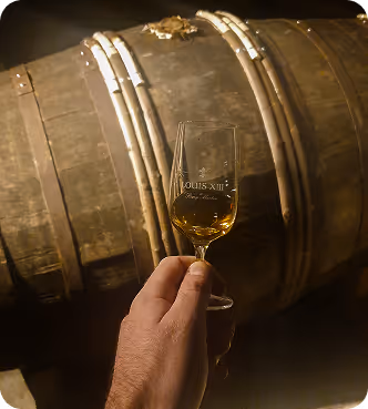Hand holding a glass of Louis XIII cognac in front of a wooden aging barrel in dim light.