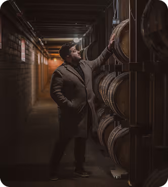 Man in coat inspecting stacked wooden barrels in a dimly lit cellar or storage room.
