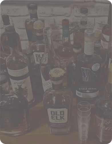 Various liquor bottles including Old Elk arranged on a wooden surface against a brick wall.