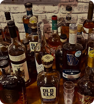Various bottles of whiskey and liquor on a wooden table against a brick wall background.