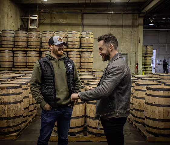 Two men smiling and fist bumping inside a warehouse filled with stacked wooden barrels.