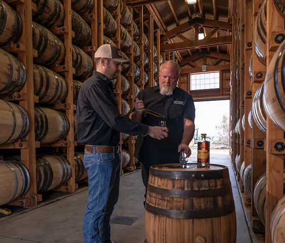 Two men in a barrel warehouse, one filling a glass with whiskey from a pipette.