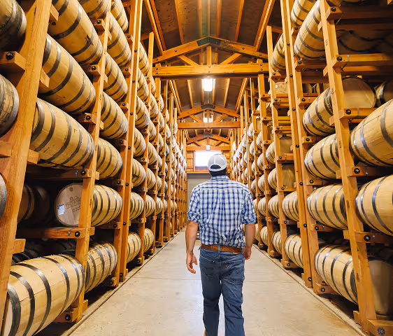 Man in plaid shirt and cap walking between wooden racks filled with stacked barrels in a warehouse.