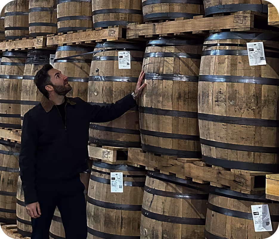 Man in dark jacket touching a wooden barrel among stacked barrels with metal bands in storage.