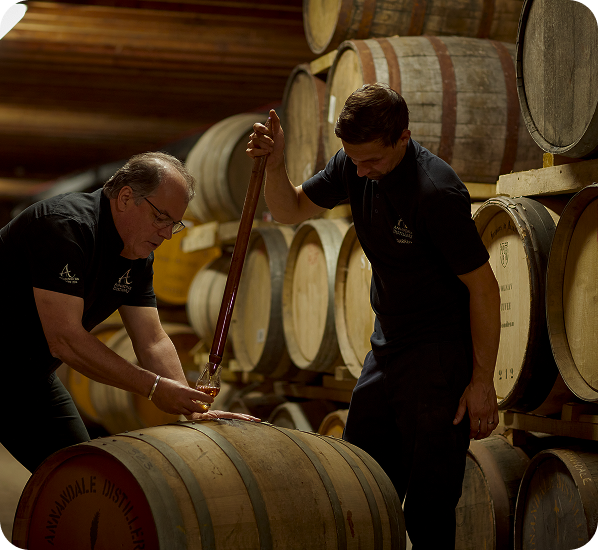 Two men sampling whiskey from large barrels in a dimly lit warehouse.