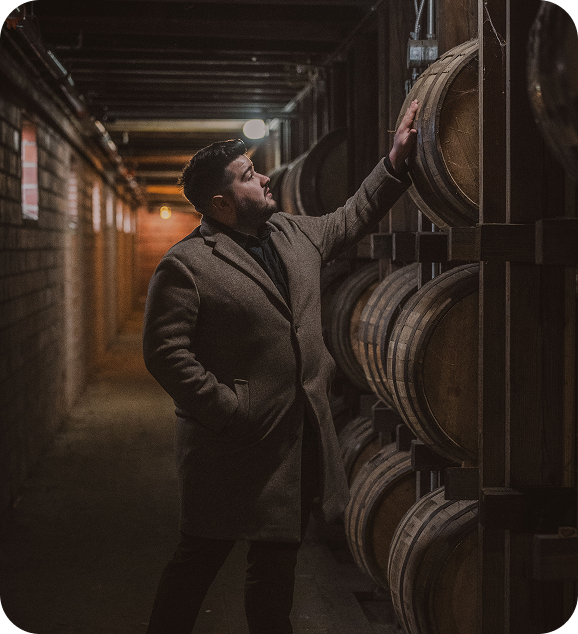 Man in a coat inspecting wooden barrels stacked in a dimly lit cellar hallway.