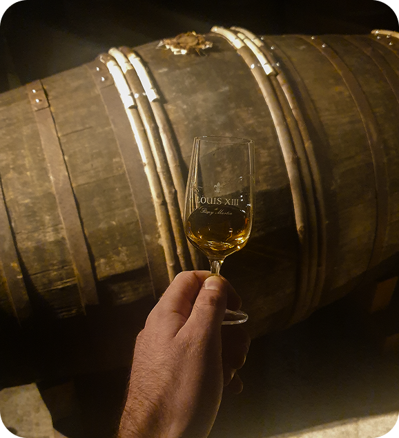 Hand holding a glass of Louis XIII cognac in front of a large wooden barrel in dim light.