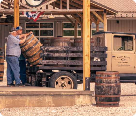 Two men loading wooden barrels onto a flatbed truck outside a distillery building.