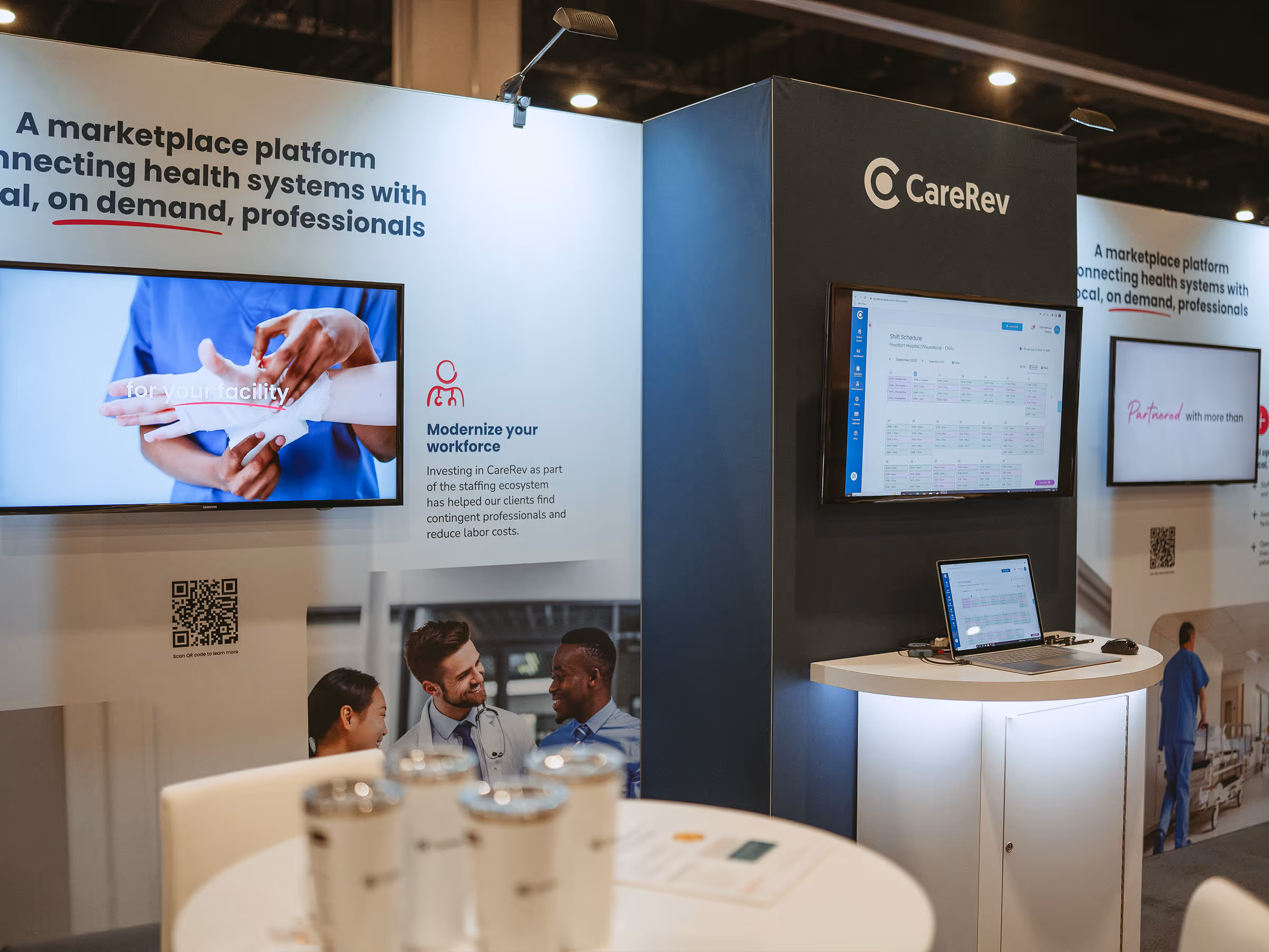 CareRev trade show booth with digital displays showing healthcare workforce scheduling and staff interacting, white and blue exhibit panels.