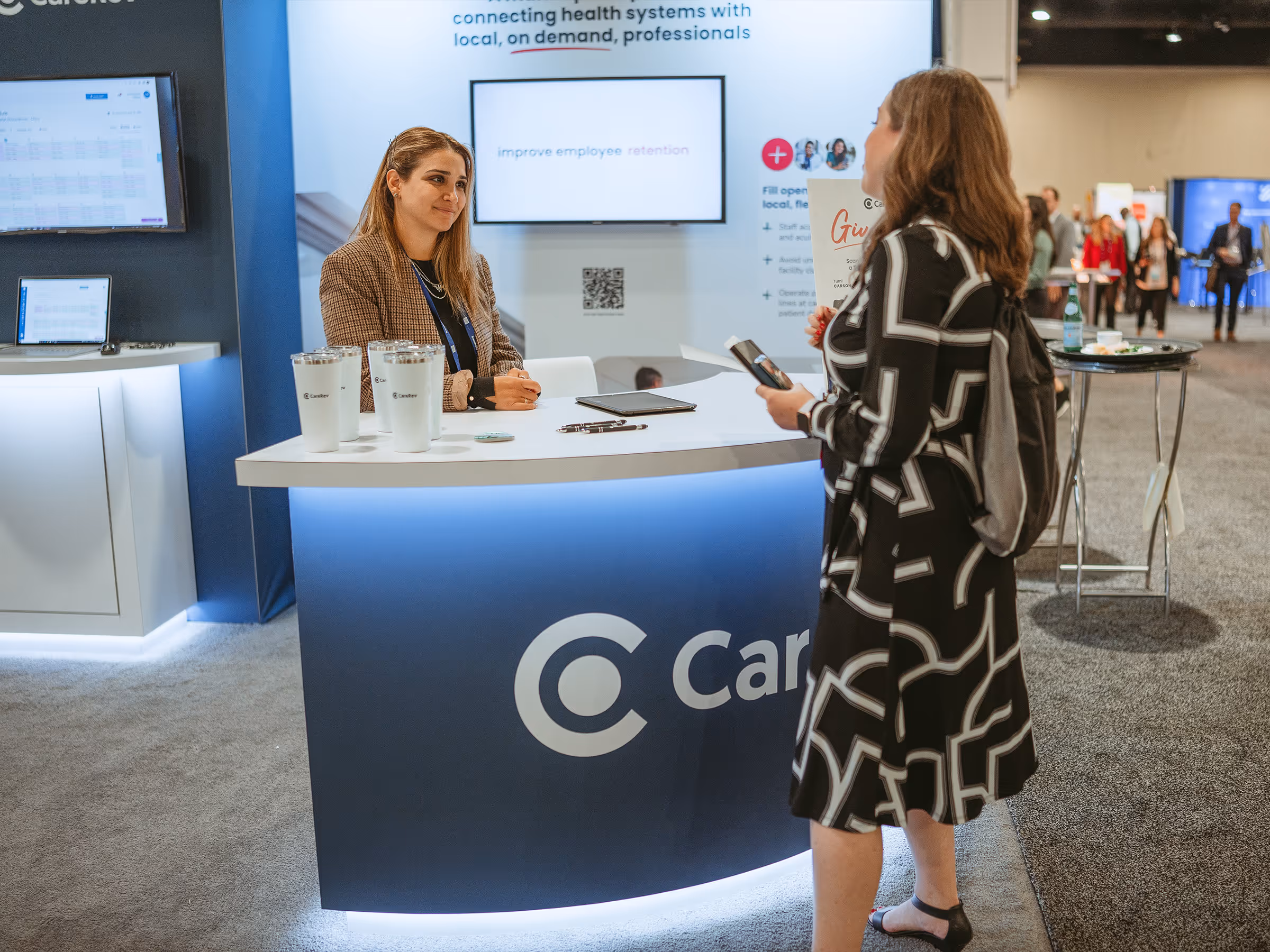 Woman with a backpack holding a phone and paper talking to a seated woman behind a CareRev booth at a conference.