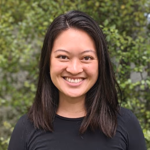 Smiling woman with straight dark hair wearing a black top standing outdoors with green leafy background.