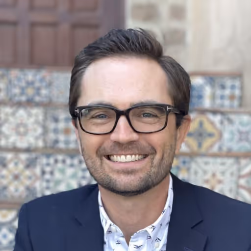 Smiling man with glasses wearing a navy blazer and patterned white shirt, standing in front of decorative tiled steps.
