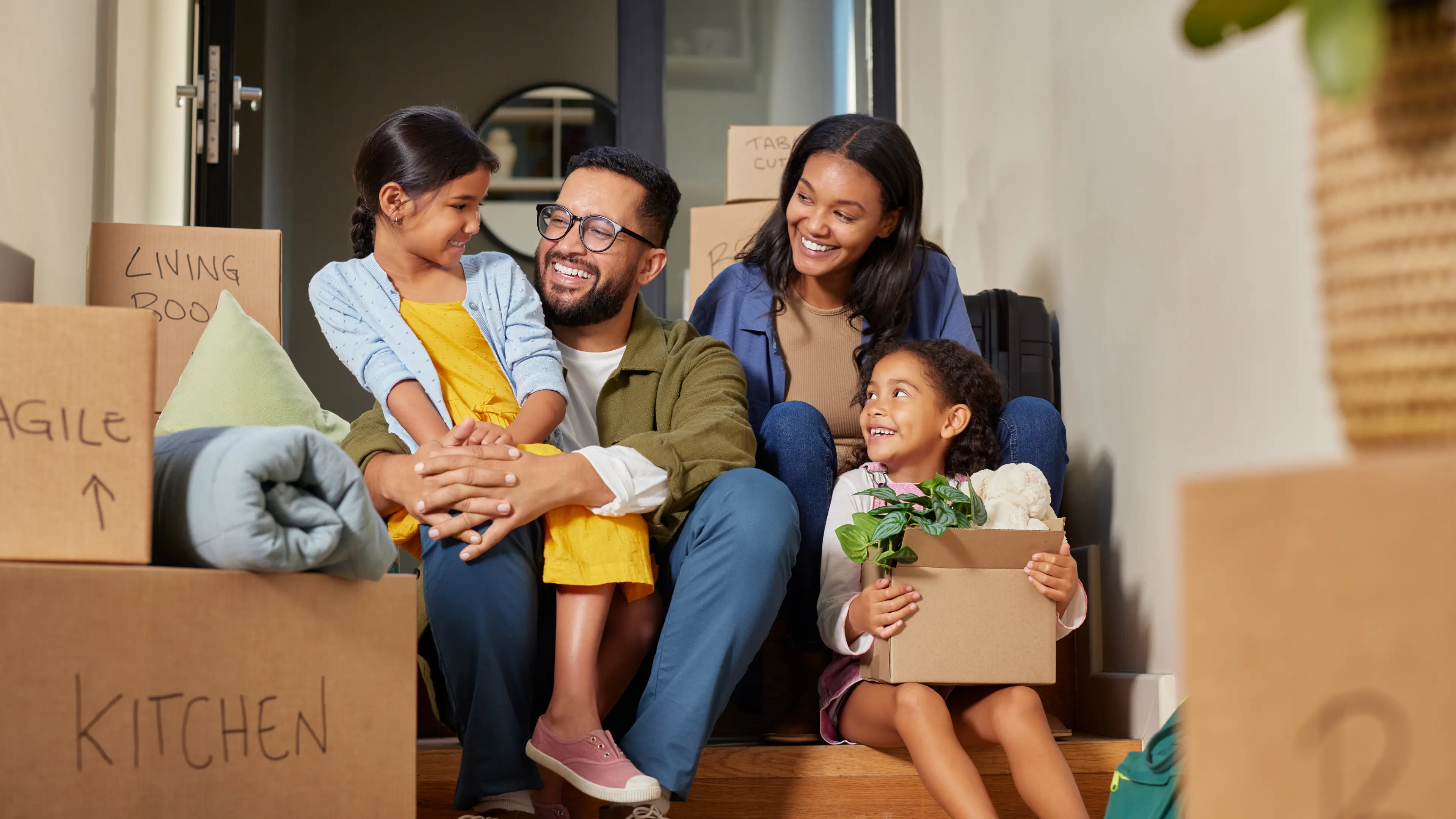 Happy family of four sitting on stairs surrounded by moving boxes labeled for different rooms.