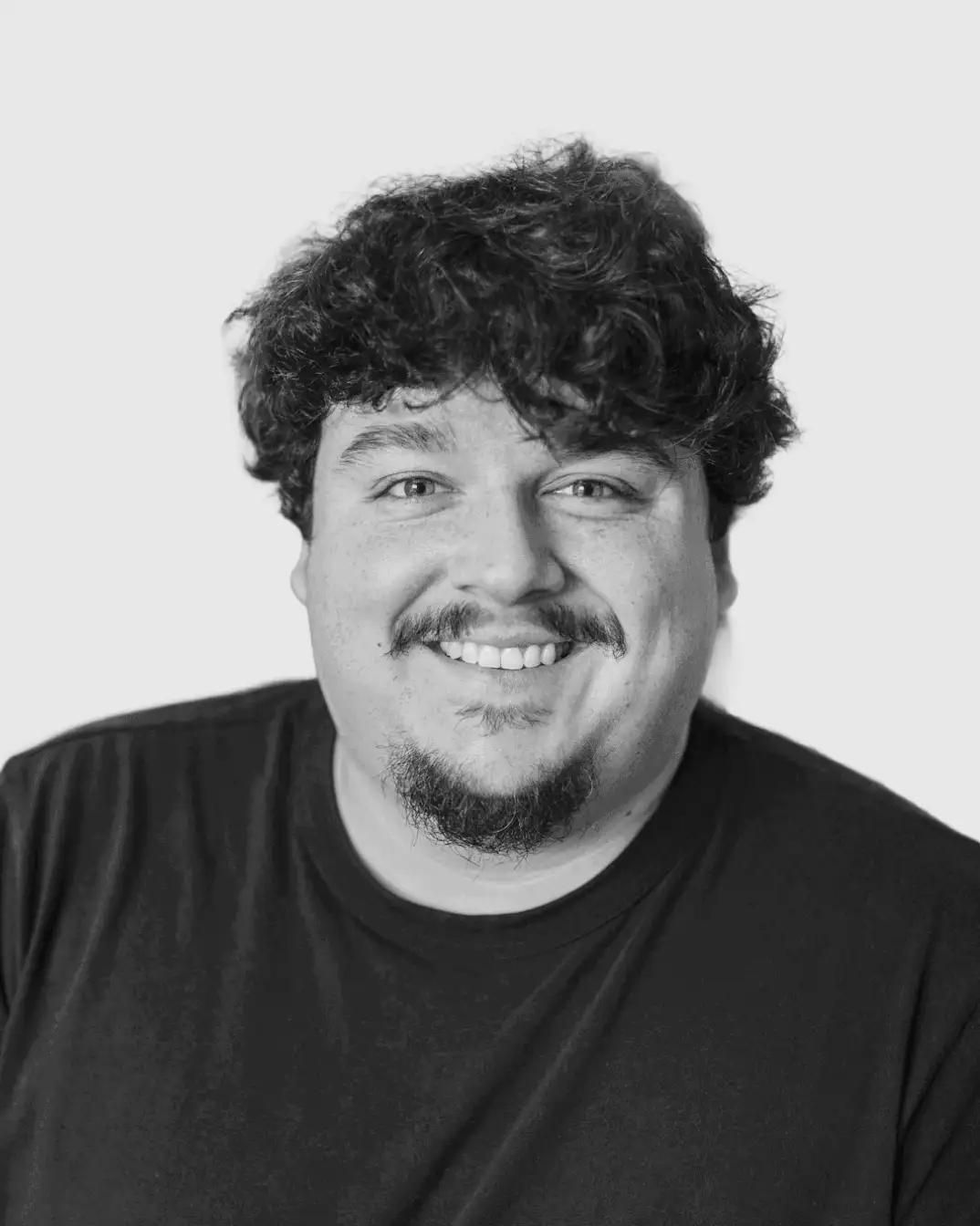 Smiling man with curly hair, a goatee, and a mustache wearing a black shirt against a plain background.