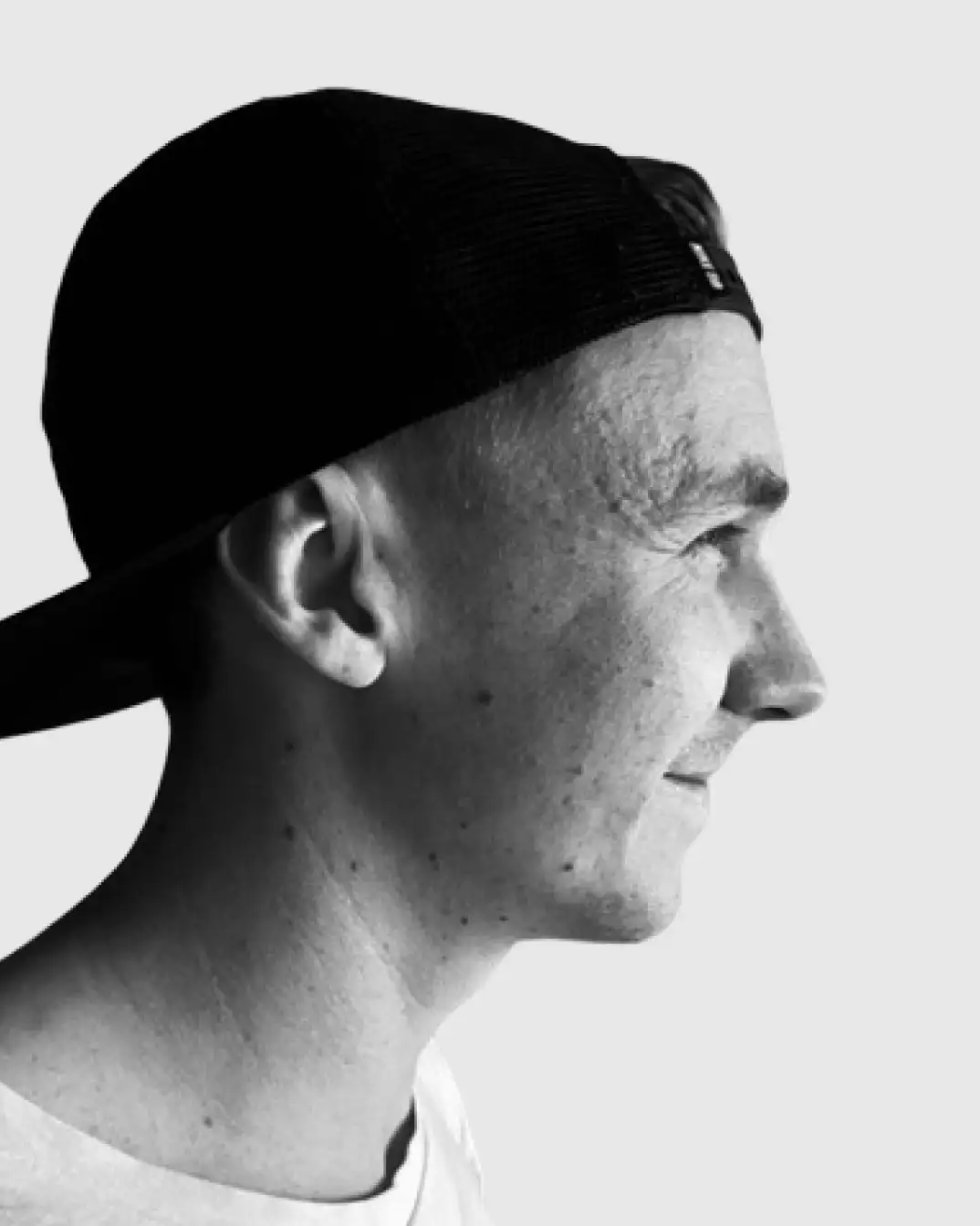 Black and white profile of a man wearing a backward black cap and a light-colored shirt, smiling slightly.