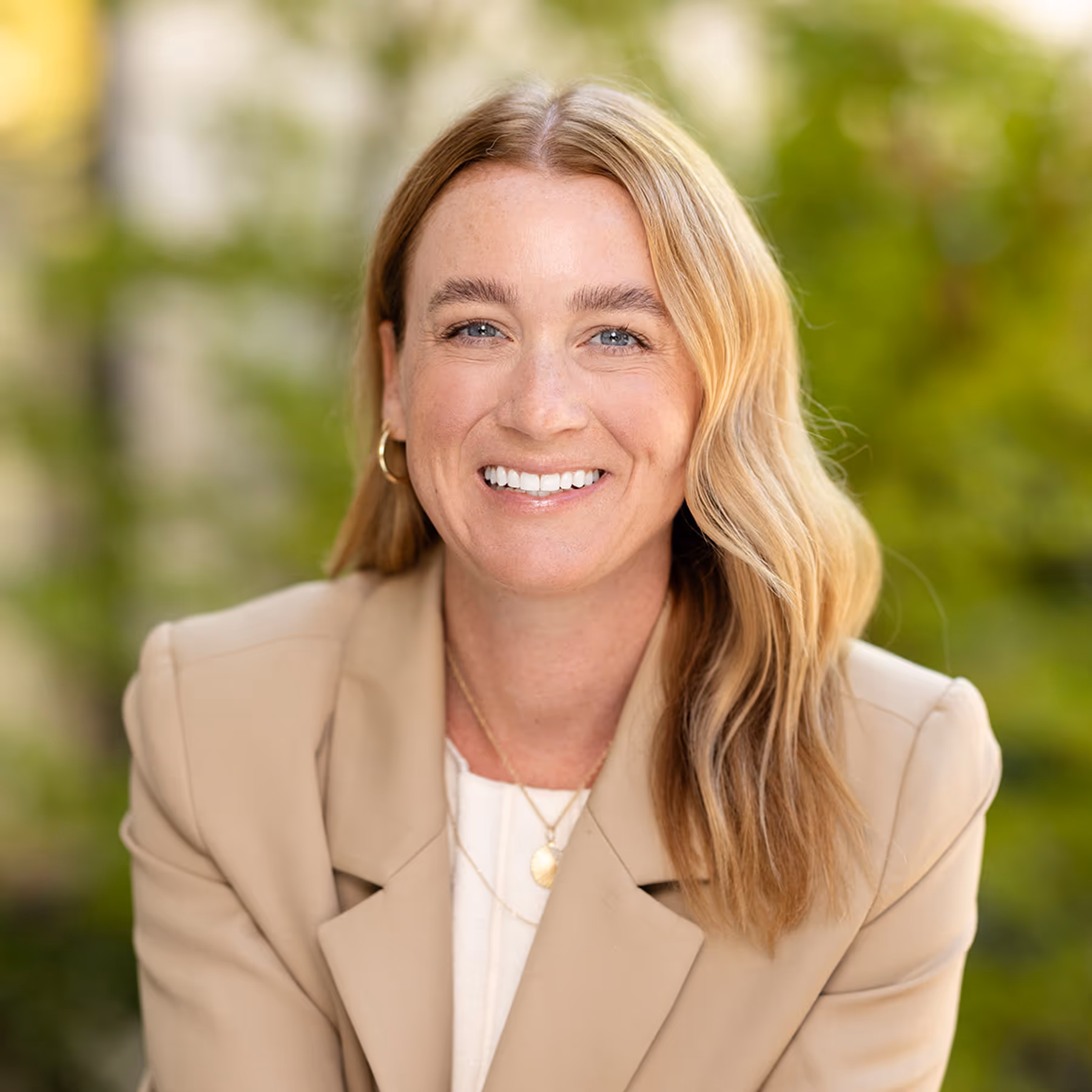 Smiling woman with blonde hair wearing a beige blazer and gold necklace in an outdoor setting.