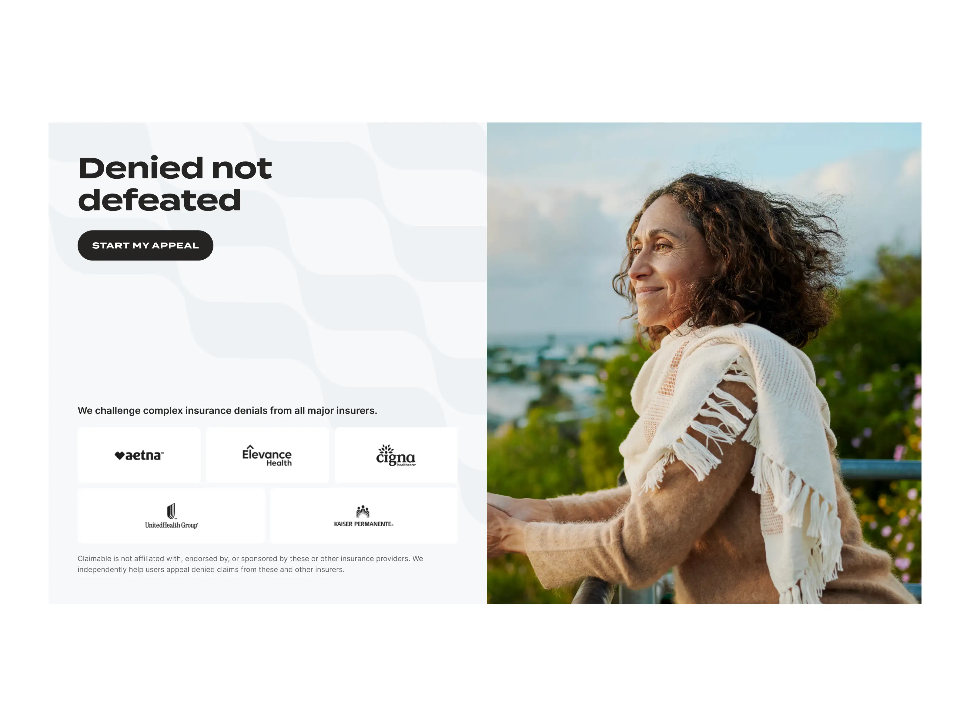 Smiling woman with curly hair wearing a beige sweater and white shawl, leaning on a railing with greenery and a cloudy sky in the background.