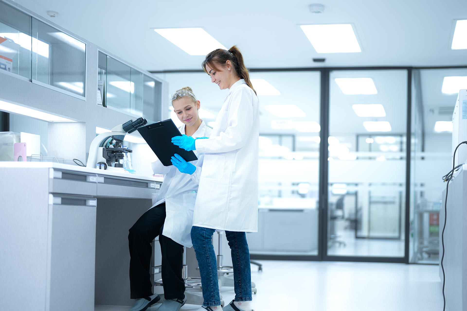 Two female scientists in white lab coats and blue gloves reviewing a clipboard in a bright laboratory with a microscope on the counter.