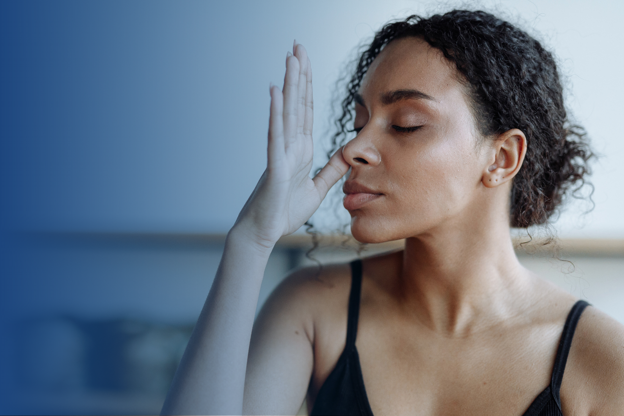 Weiblich gelesene BIPOC Person atmet bewusst mit geschlossenen Augen. Sie hält sich ein Nasenloch zu.
