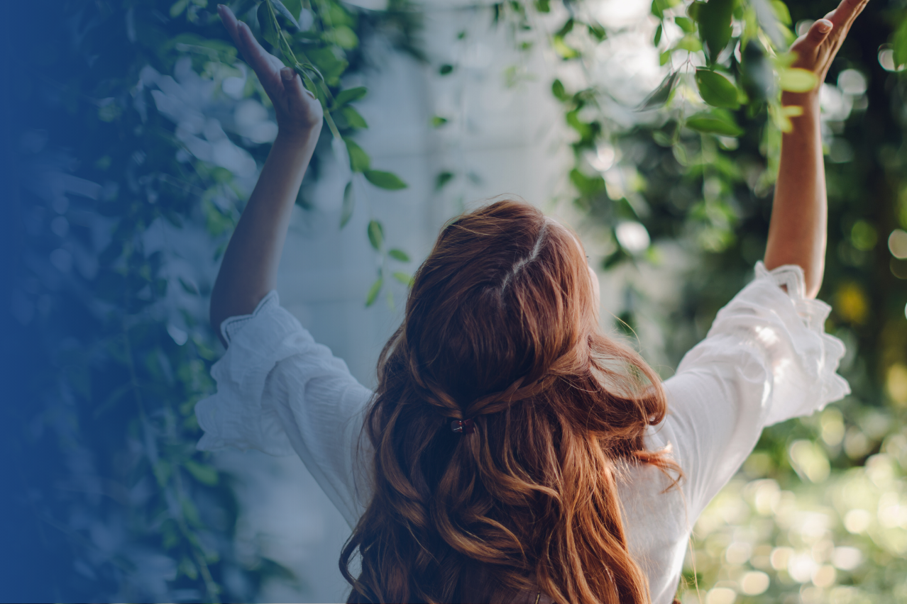 Eine weiblich gelesene Person mit langen Haaren steht mit dem Rücken zum Fotografen in der Natur - Kopf und Hände hält sie nach oben in die Luft. 