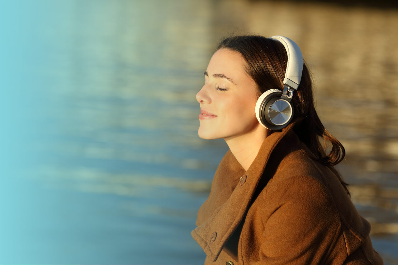 Eine weiblich gelesene Person mit langen Haaren sitzt mit Kopfhörern auf am See, hat die Augen geschlossen und hört lächelnd einer Meditation zu.