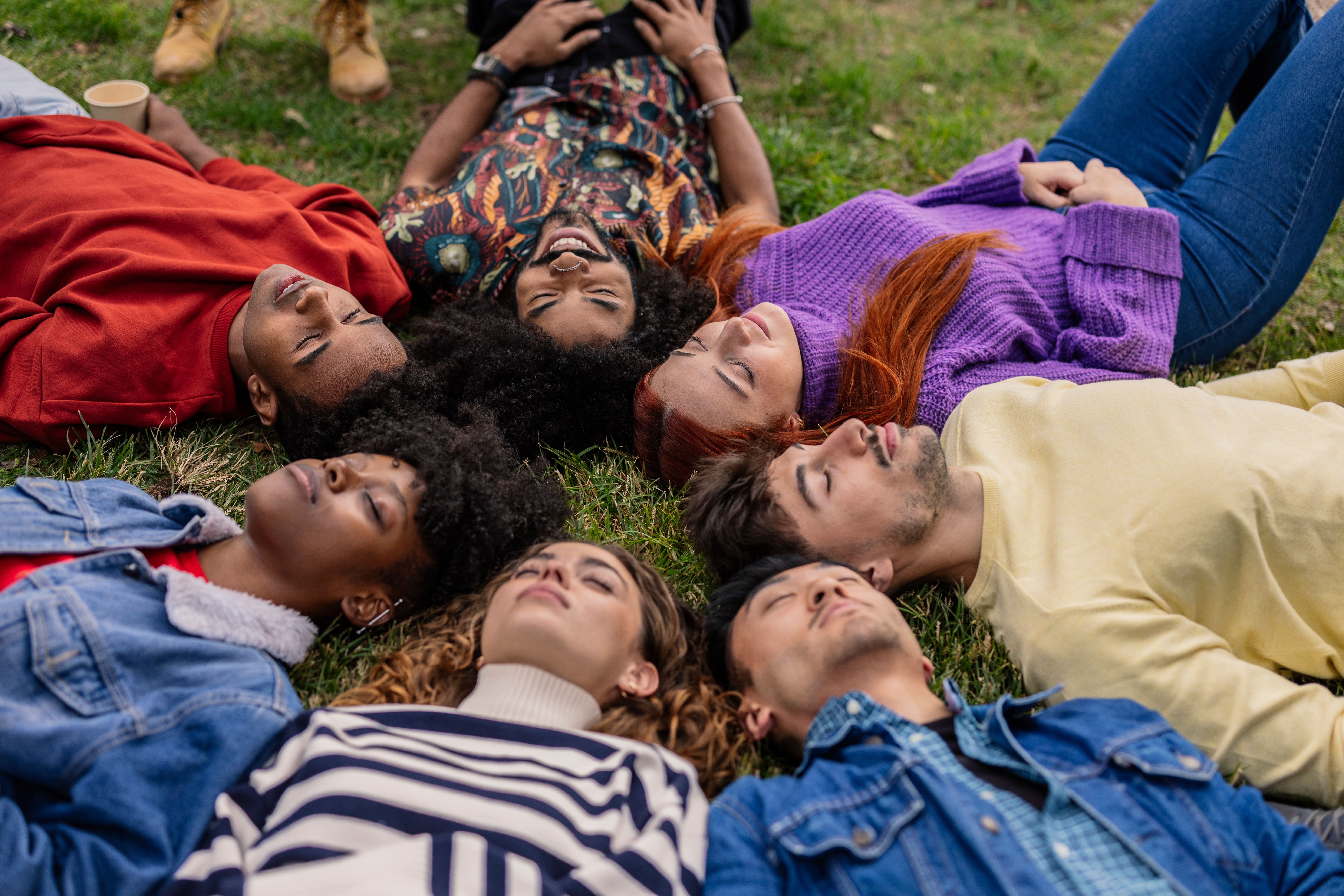 A diverse group of seven people lie in a circle on the gras, heads together, eyes closed, consciously breathing.