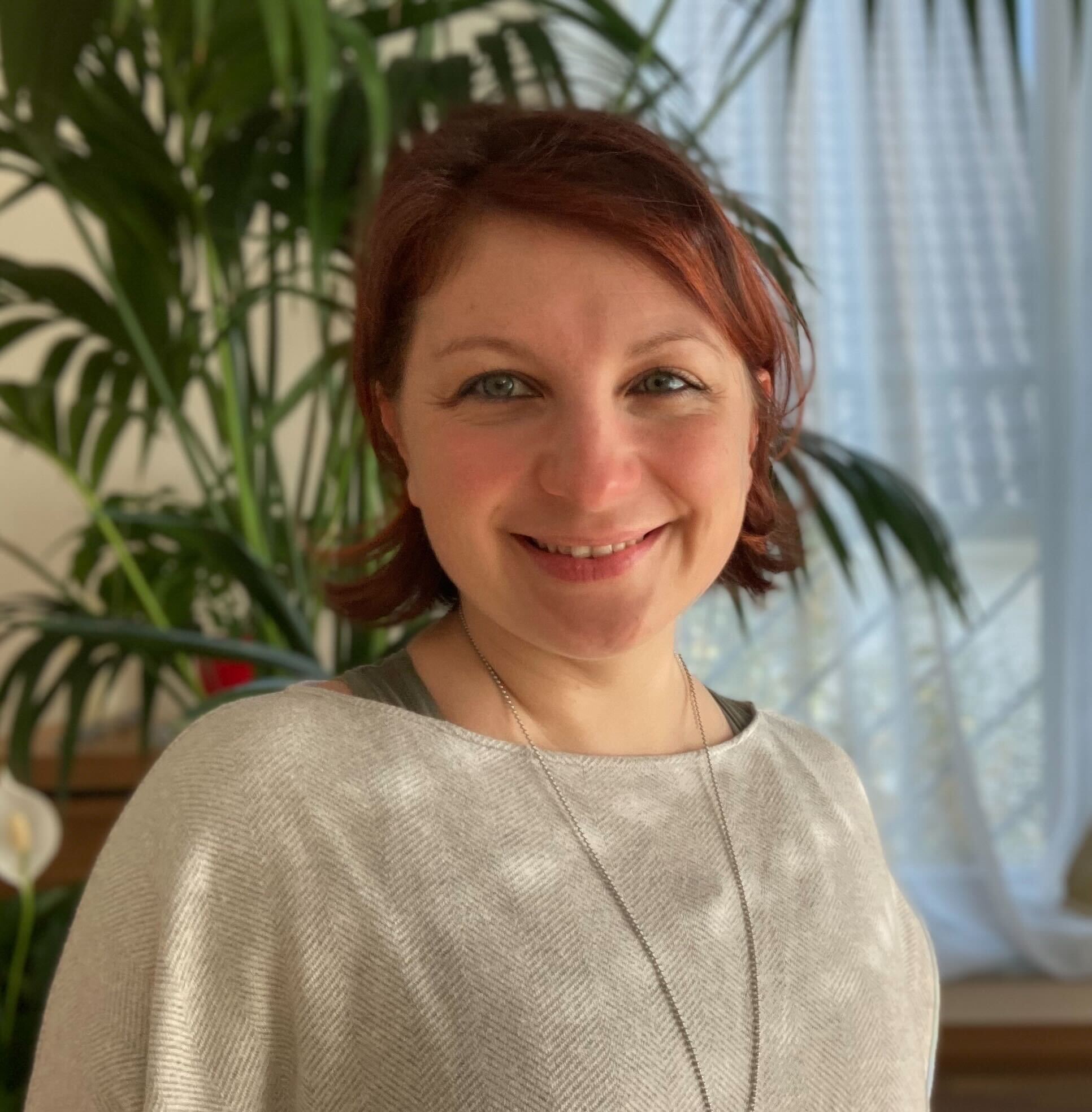 Head Shot von Lucia, die herzlich in die Kamera lacht. Im Hintergrund steht eine Pflanze.