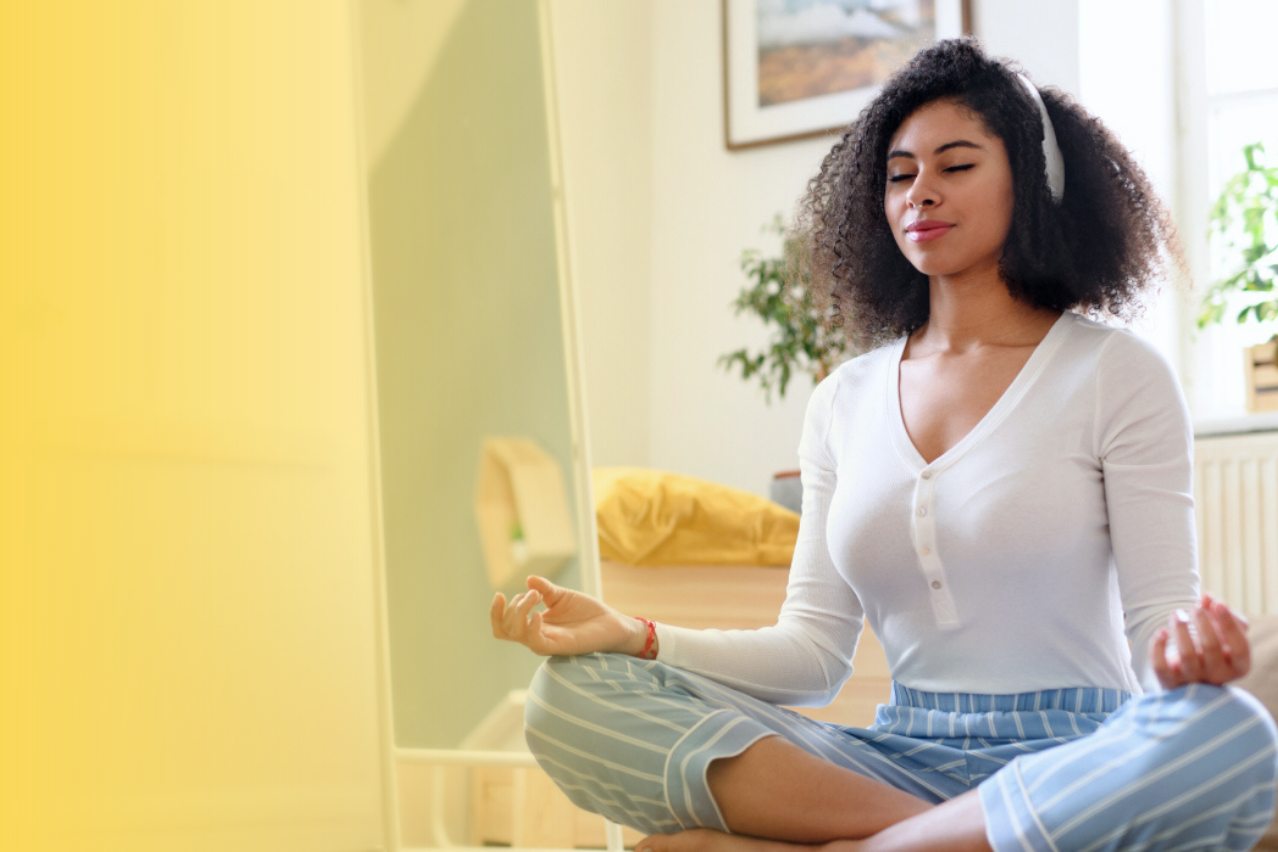 Eine weiblich gelesene Person of Color sitzt auf dem Boden, im Schneidersitz mit geschlossenen Augen, und meditiert.