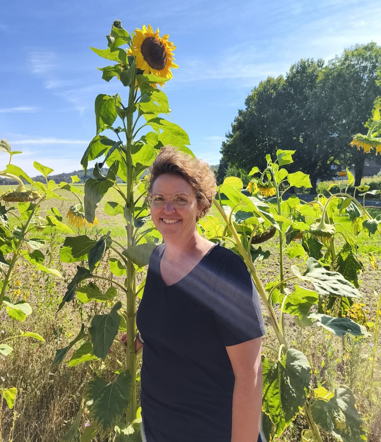 Foto von Bianca, die vor einem Feld voller Sonnenblumen steht. Sie trägt ein schwarzes T-Shirt und kurze Haare. Sie lacht offen in die Kamera.