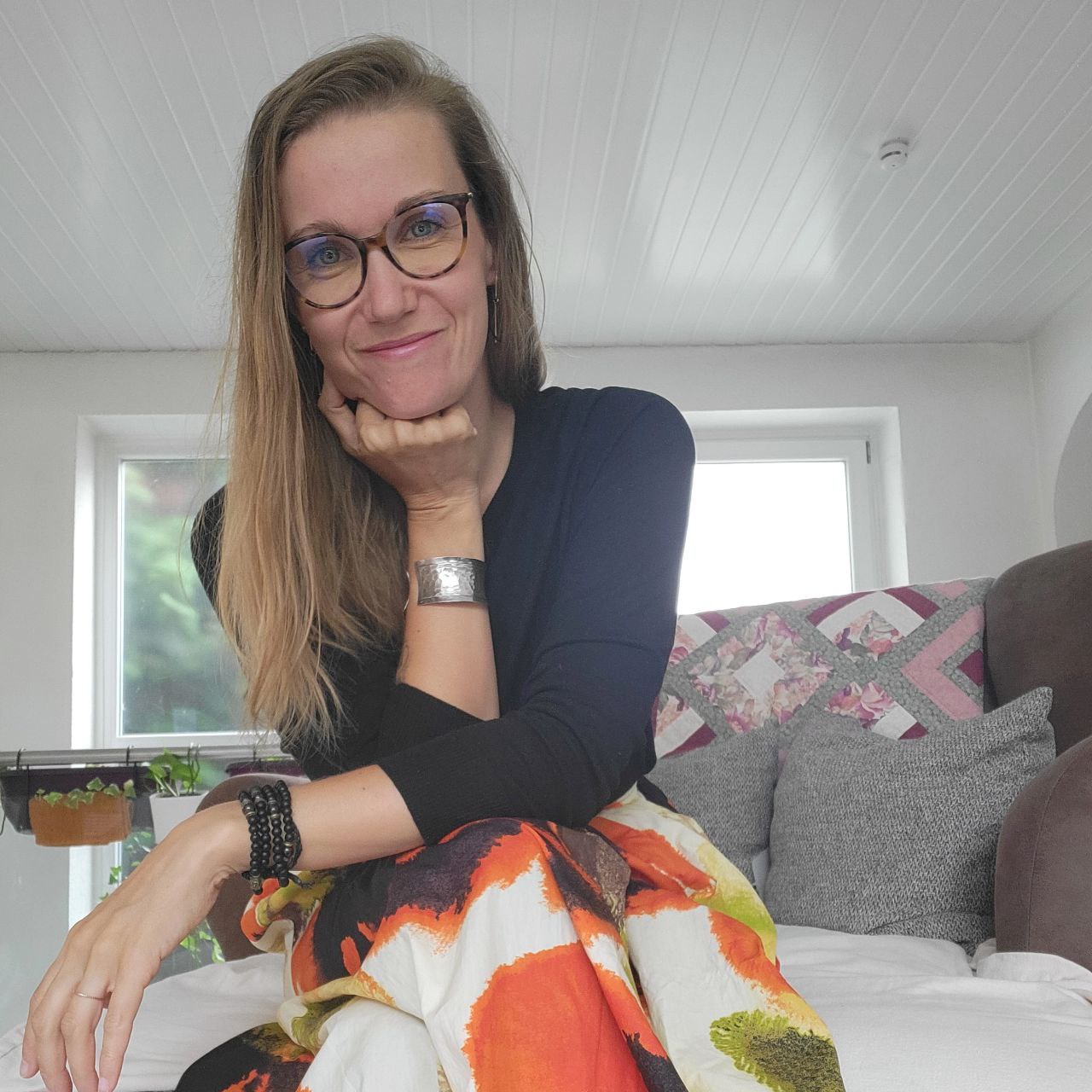 Foto von Jenni, die auf ihrem Sofa sitzt, die Beine überschlagen und ihren Kopf auf einem Arm abgestützt, leicht nach vorne gelehnt, auf dem Knie abgestützt. Sie trägt ein dunkles Shirt und eine Brille und hat blonde, lange Haare, die ihr offen über die Schulter fallen.