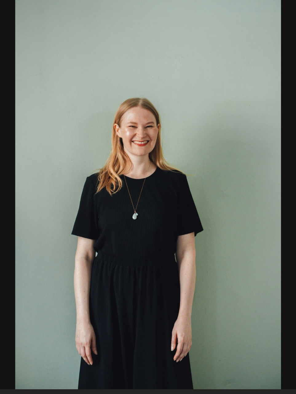Foto von Lisa, die vor einem blass grünen Hintergrund steht. Sie trägt schwarze Kleidung und hat lange blonde Haare, die ihr offen über die Schultern fallen. Sie lacht in die Kamera. 
