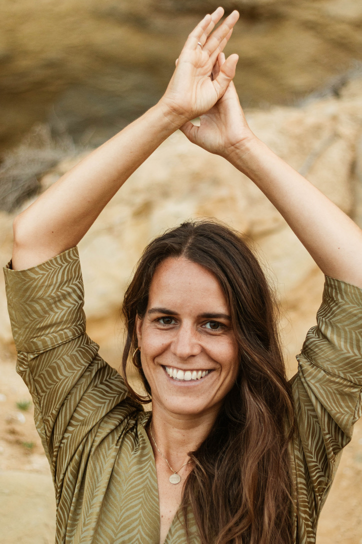 Foto von Sonja, die in der Natur steht. Sie trägt ein grünes Shirt und hat lange, braune Haare, die offen über ihre Schulter fallen. Sie hält die Arme wie in einer tanzenden Bewegung ausgestreckt über den Kopf.