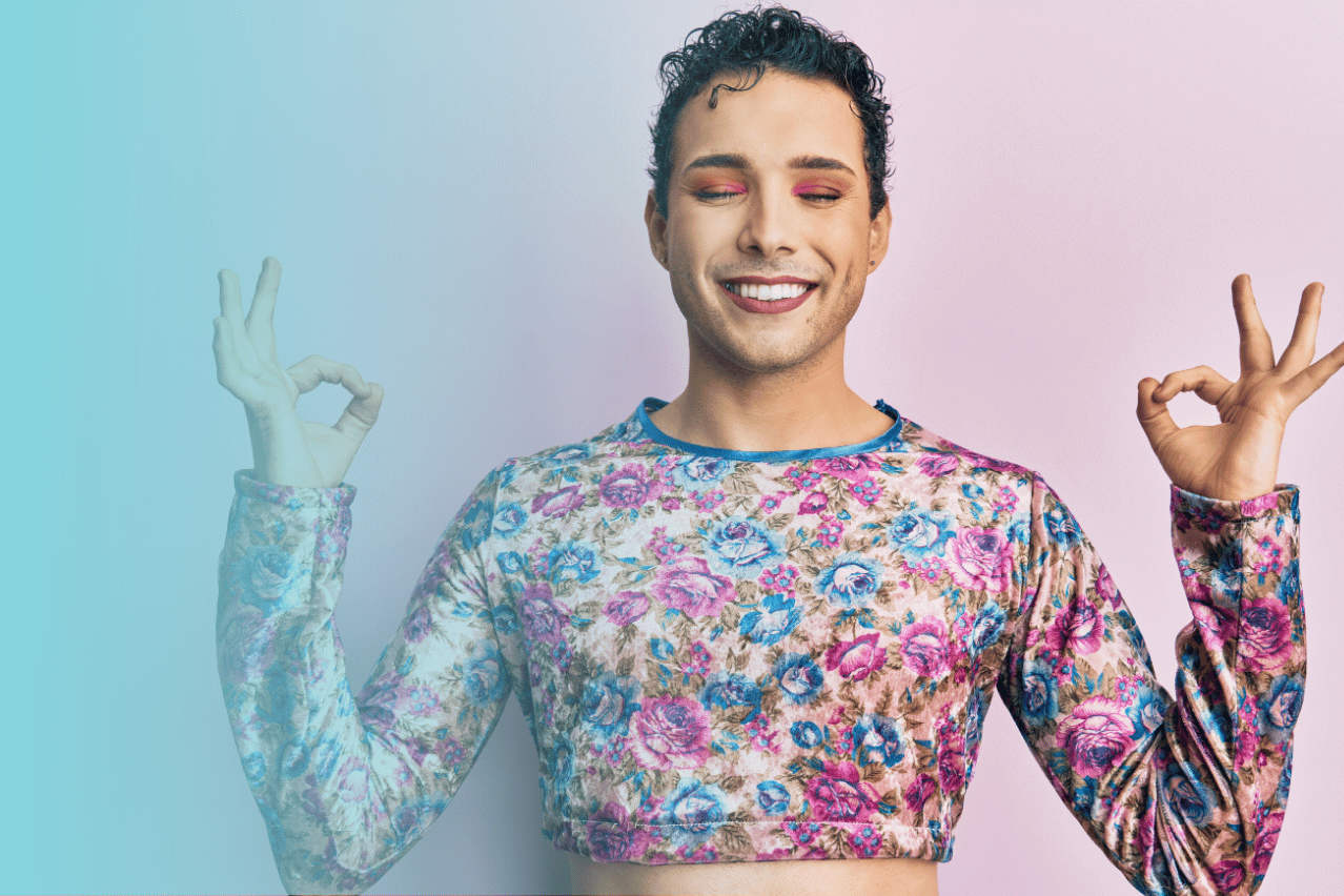 Eine nicht binäre Person steht in buntem Shirt und schönem Make-up vor einer rosa-farbenen Wand. Sie hat die Augen geschlossen und hält die Hände in Meditationshaltung. Sie lächelt und atmet bewusst.