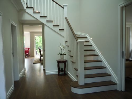 A modern, minimalist interior featuring a sleek wooden staircase with vertical slats acting as a railing and divider. Two geometric pendant lights hang above, illuminating the warm wood tones. In the background, a dining area with a wooden table and chairs is visible.