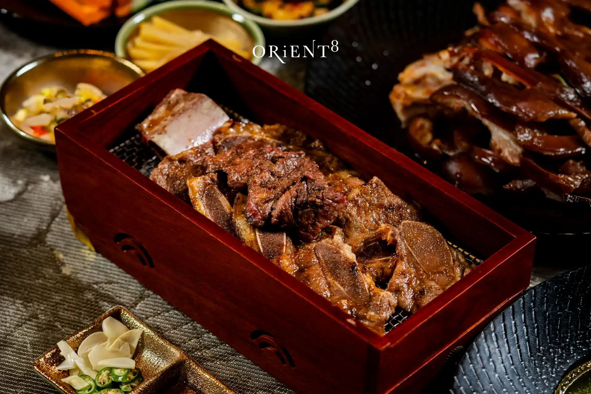 Grilled Yangnyeom Galbi short ribs in a wooden box with banchan and sauces, part of the Korean Barbecue Favourites at Orient8, Hotel Mulia Senayan Jakarta.