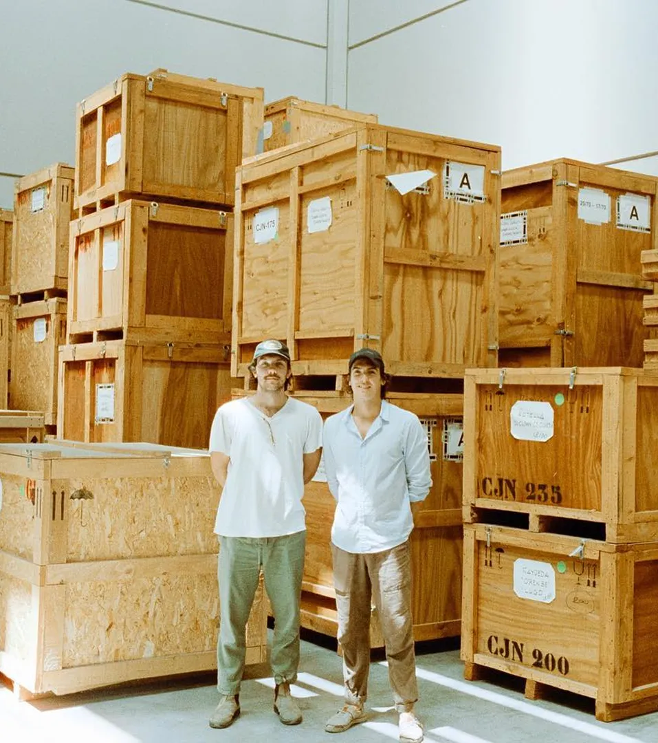Two men wearing casual clothes and caps standing in front of large stacked wooden crates inside a warehouse.