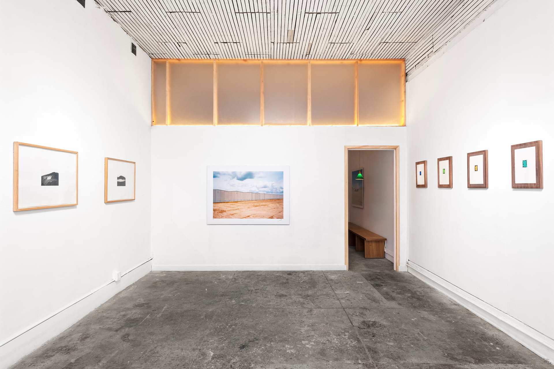 Minimalist art gallery room with white walls, concrete floor, and framed photographs; one large photo centered, two small ones on the right wall, and one on the left wall, with lit translucent panels near the ceiling.