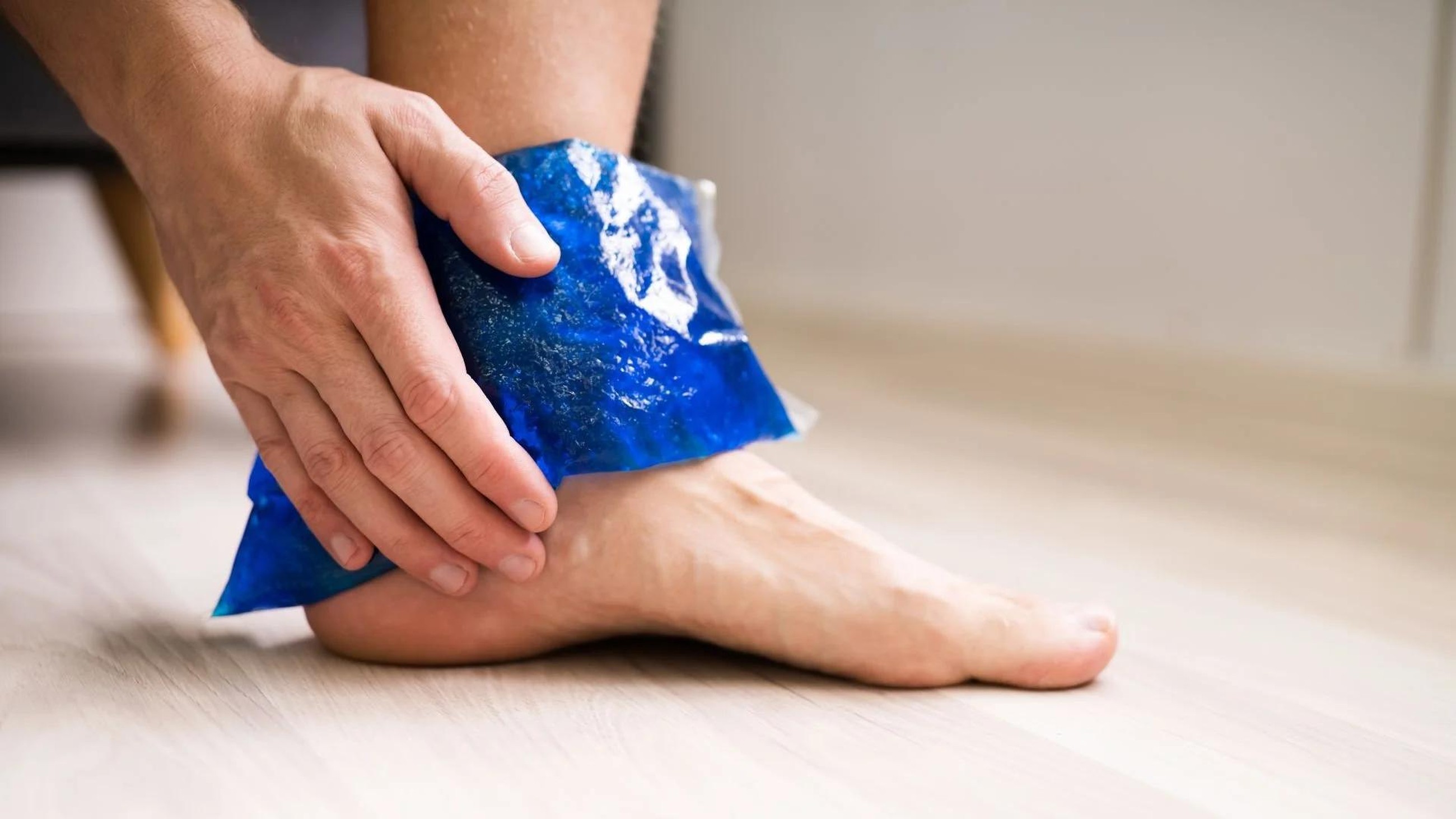 A person applying a blue ice pack to their ankle to relieve pain and swelling following an injury.