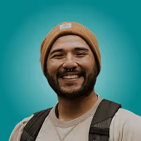 Smiling man wearing a beige beanie and a backpack against a teal background.