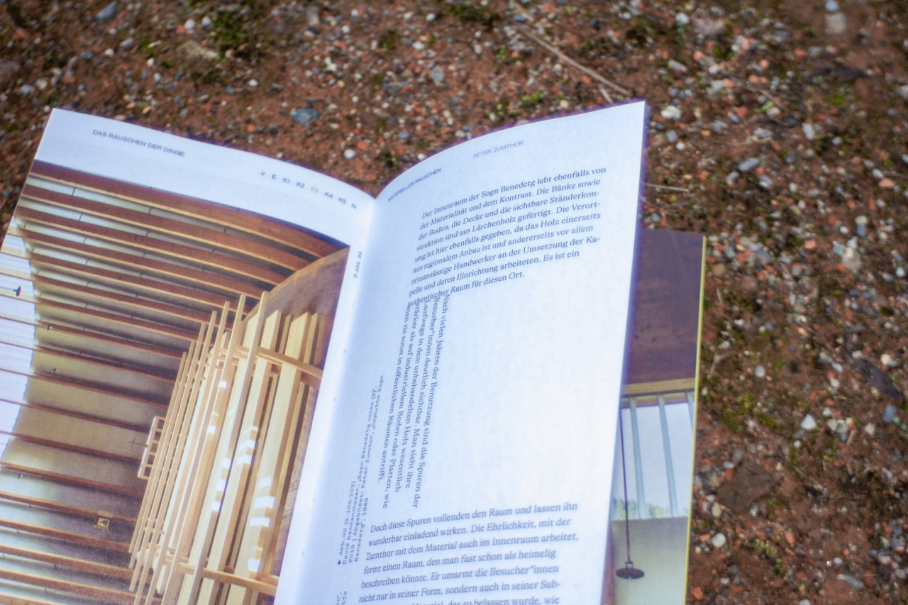 Foto eines aufgeschlagenen Buchs mit Architekturtext und Bild, auf einem erdigen, mit Moos bedeckten Boden liegend.