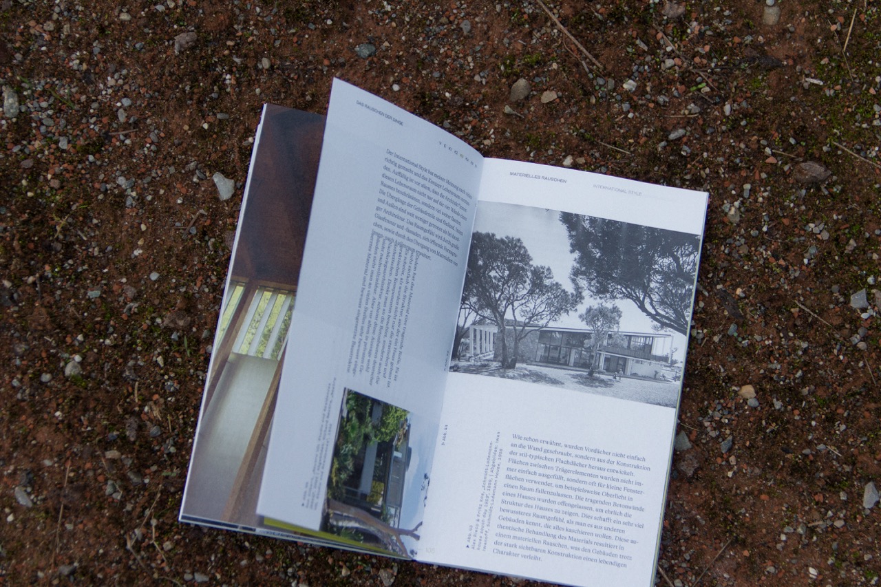 Offenes Architekturmagazin mit Text und Schwarz-Weiss-Bild eines modernen Hauses liegt auf steinigem, erdigen Boden.