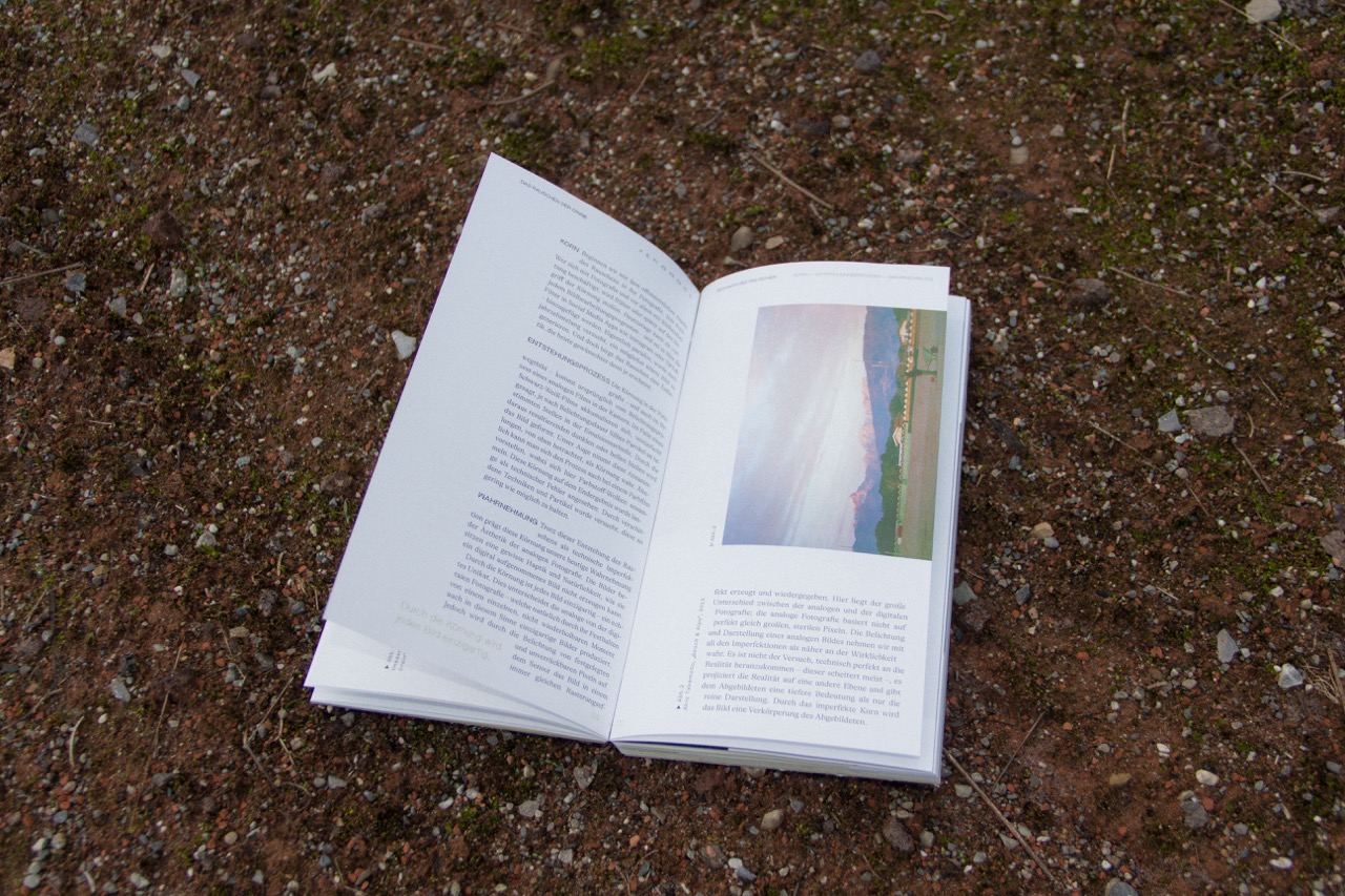 Ein aufgeschlagenes Buch mit Text und einem Landschaftsfoto liegt auf einem erdigen und steinigen Boden.