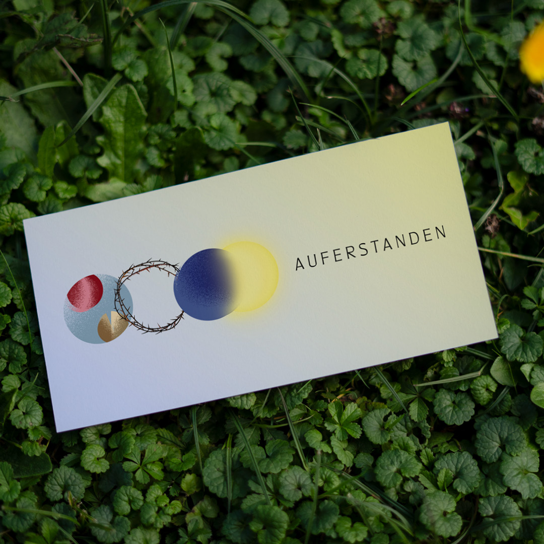 Weiße Karte mit stilisierten Kreisen, Dornenkrone und dem Text ‚AUFERSTANDEN‘, auf grünem Gras liegend.