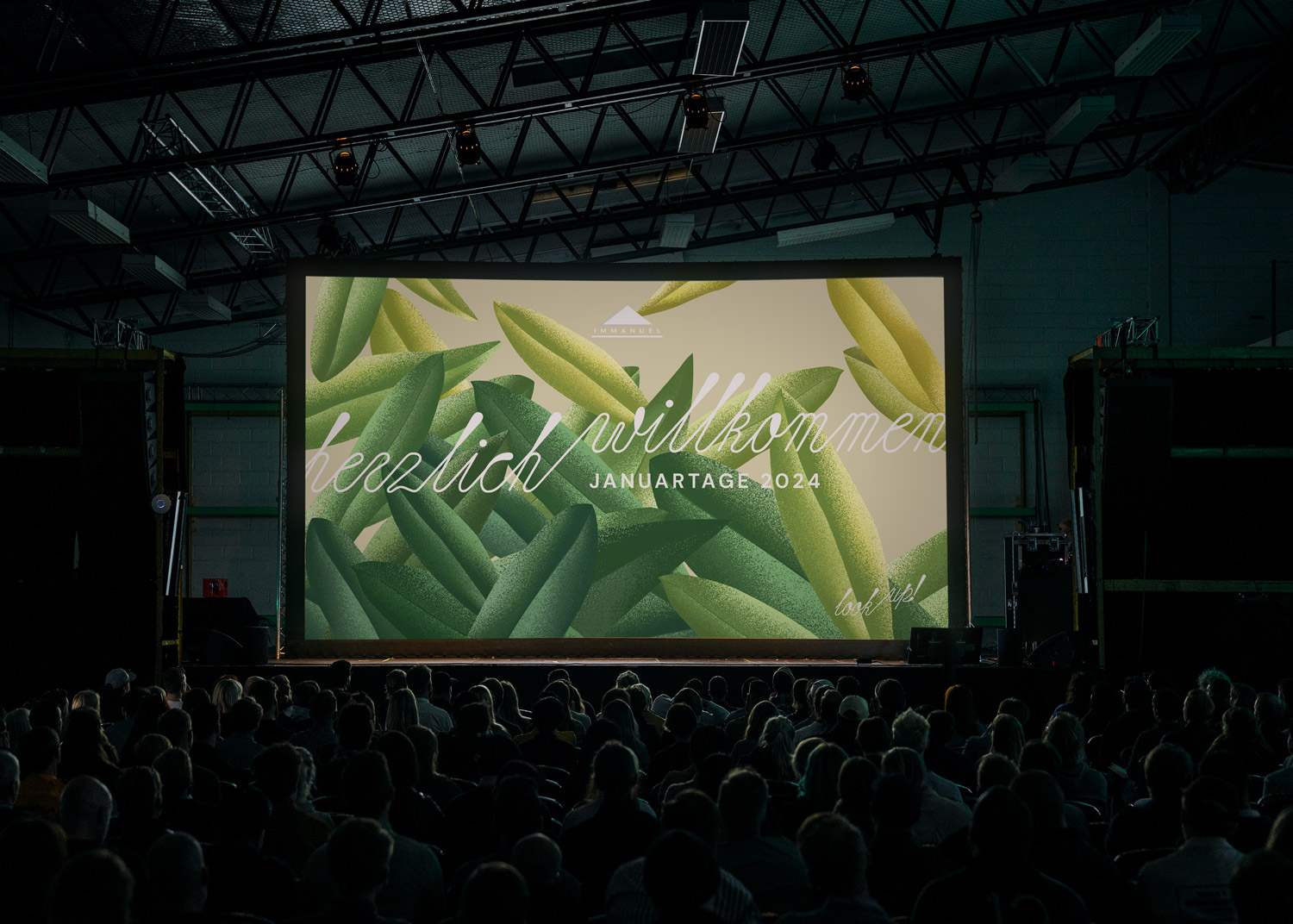 Publikum in einer dunklen Halle vor einer grossen Leinwand mit einer grünen Blattgrafik und dem Text «herzlich willkommen Januartage 2024».